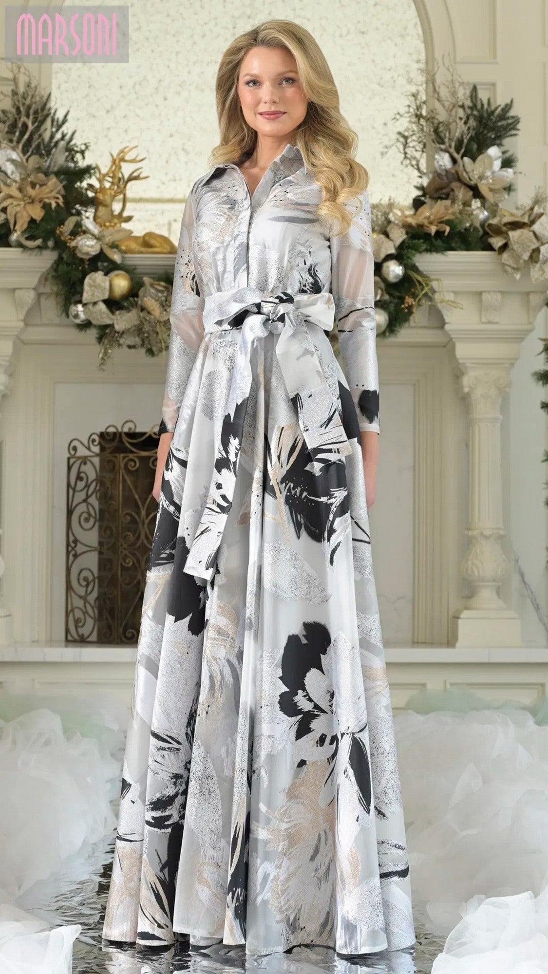A woman stands indoors in front of a decorated fireplace, wearing the Marsoni MV1367 long formal dress in Silver Multi with a floral organza jacquard pattern, belted waist, and A-line silhouette.